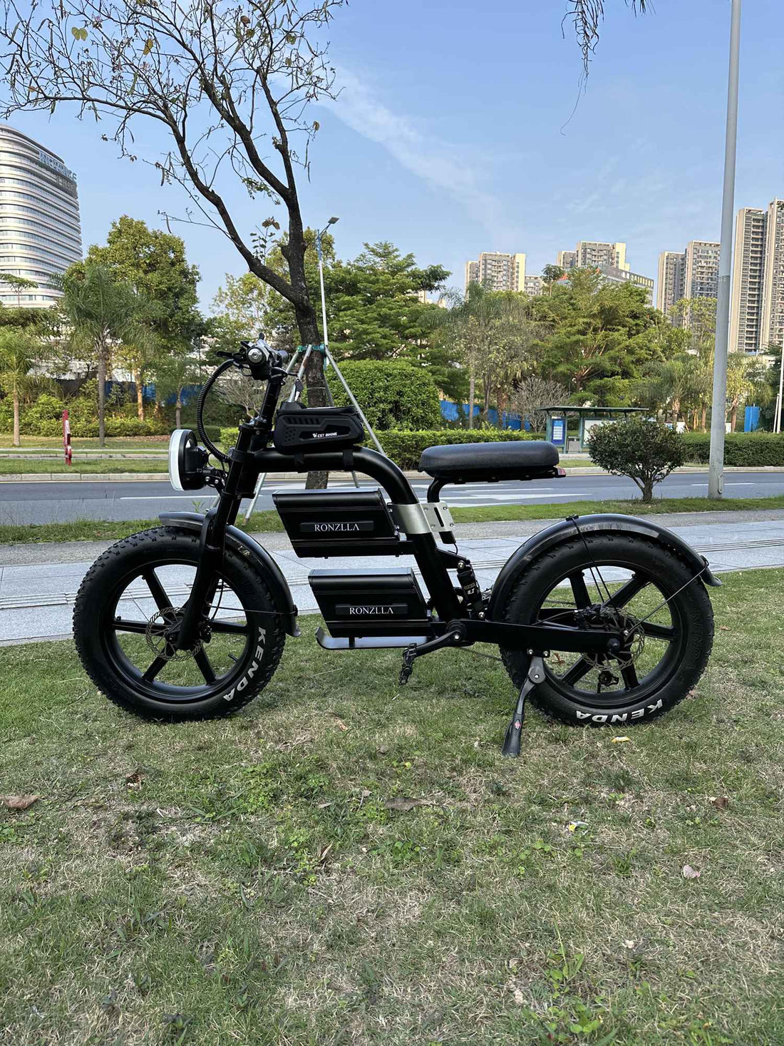RONZLLA E20 Plus black electric dirt bike with KENDA fat tires and dual battery setup parked on the grass at an urban park.