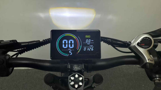 Close-up shot of a RONZLLA E20 e-bike handlebar featuring a bright, digital LCD display screen that shows zero speed, an illuminated headlight beam on the wall, and Shimano gear shifters on the sides.