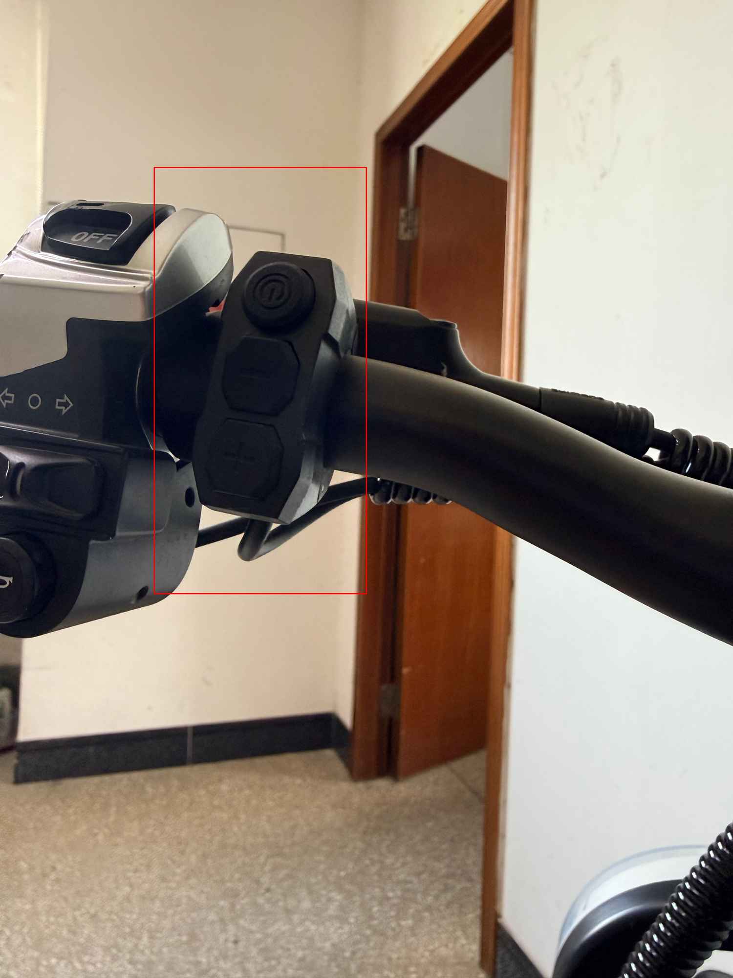A close-up photo of the black handlebar on a RONZLLA E20 Plus electric bicycle. A red box highlights a vertical three-button control switch, showing the power button, menu button, and gear adjustment button from top to bottom. The horn and turn signal control unit is visible on the left.