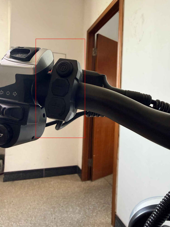 A close-up photo of the black handlebar on a RONZLLA E20 Plus electric bicycle. A red box highlights a vertical three-button control switch, showing the power button, menu button, and gear adjustment button from top to bottom. The horn and turn signal control unit is visible on the left.