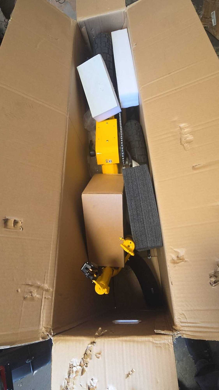An open cardboard shipping box showing the interior protective foam packaging and components of a yellow wholesale used electric bike inside a US warehouse.